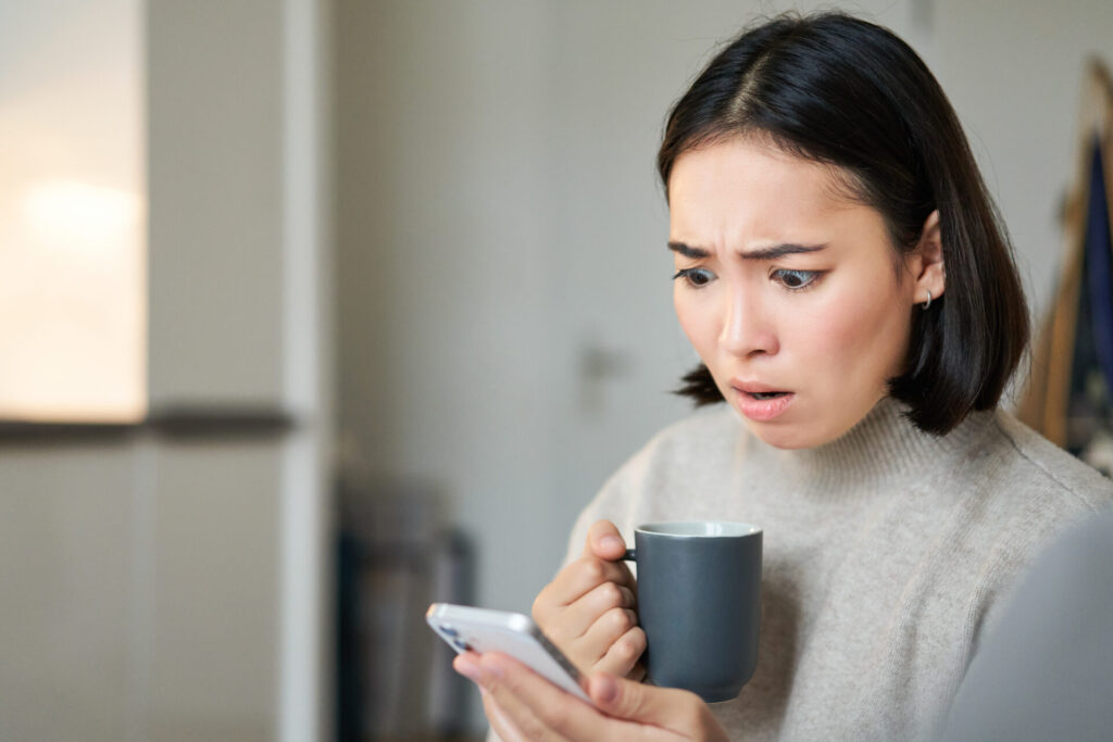 カップを手に持ち、携帯電話の画面にショックを受けながら、スマートフォンアプリで気になるニュースを読んでいるアジア人の女の子のクローズアップ
