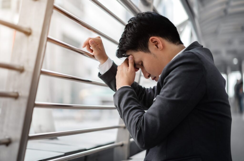 頭を抱えて手すりにもたれかかり、仕事のミスに落ち込んでいるスーツ姿の日本人ビジネスマンが屋外通路でうなだれている様子